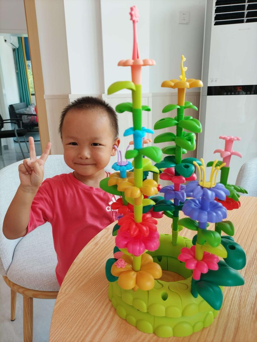 孩子坐在桌前举着剪刀手，旁边是照老师模板拼好的彩色花树玩具