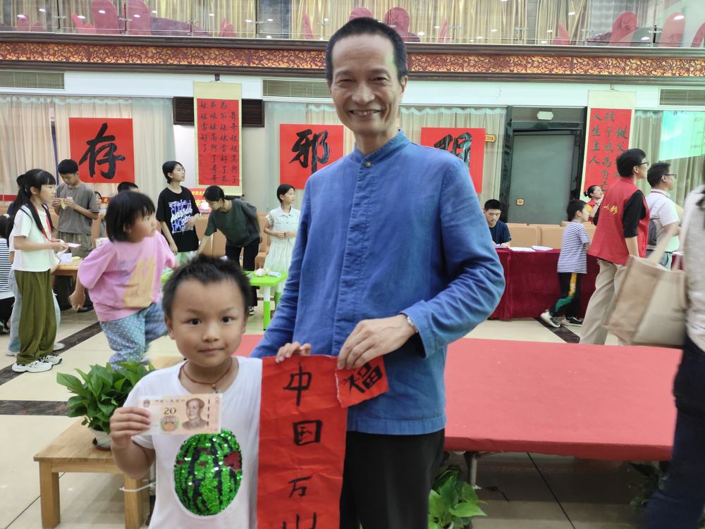 孩子与一位身着中式布衫的师者在书法活动现场合影，手持中国万岁与福字